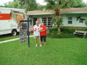Three people stand in front of a house with a sold-for sign.