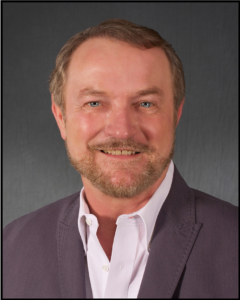 Smiling middle-aged man in a suit jacket and white shirt against a gray background.
